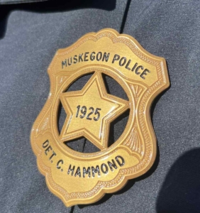 Golden police badge with "Muskegon Police," "1925," and "Det. C. Hammond" inscribed. It rests on a dark fabric, conveying authority and tradition.