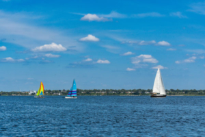 Two colorful sailboats with bright sails and a white sailboat glide across Muskegon Lake under a clear sky.