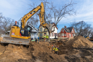 A large excavator digs a deep pit in a residential area, with workers in safety gear guiding the process. Cozy houses and bare trees are in the background.