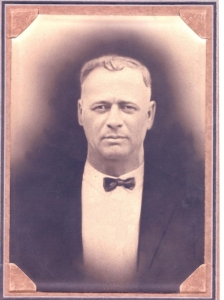 Sepia-toned portrait of a man in a suit with a bow tie, looking confidently at the camera. The vintage style evokes an early 20th-century feel.