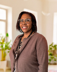 Smiling woman with glasses, wearing a brown jacket, stands in a bright room with large windows and green plants, conveying a warm, welcoming feel.