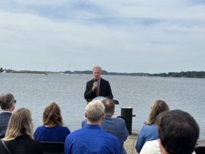 A person in a suit speaks at a podium near a calm waterfront with sailboats and distant land. Audience members sit in the foreground.