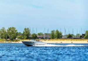 A white speedboat glides swiftly across Muskegon Lake, leaving a white wake behind. In the background, a sandy shoreline with trees and sailboats.