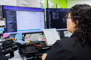 A woman with curly hair, seen from the side, works at a desk with dual monitors displaying detailed documents and data against a colorful office backdrop.