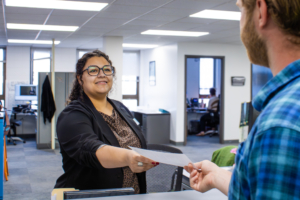 A woman in an office hands a document to a man. She is smiling, wearing glasses and a blazer. The office is bright with people working in the background.