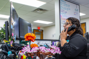 A woman in an office talks on the phone at her desk. Dual monitors display work, while colorful flowers and notes add a vibrant touch. A whiteboard is in the background.