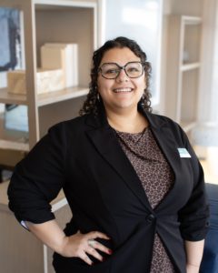 Smiling individual with curly hair and glasses, wearing a black blazer over a patterned top, stands confidently in a bright office setting.