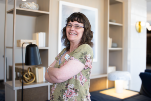 Smiling woman with glasses and floral blouse stands confidently with arms crossed in a cozy living room, featuring warm lighting and bookshelves.