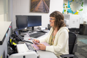 A woman with glasses smiles while working at a desk, holding papers. She is surrounded by computers and office equipment, creating a cheerful, productive atmosphere.