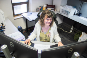 A woman with glasses sits at a desk, smiling at two computer monitors. The office is cluttered with papers, creating a busy and positive work atmosphere.