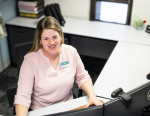A woman in a pink blouse smiles while seated at a desk with a computer in an office. The setting is bright and organized, conveying a positive work atmosphere.