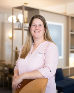 A woman with long hair, wearing a pink shirt, smiles warmly while leaning on a chair in a modern, well-lit room with shelves and soft lighting.