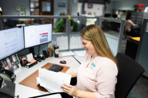 A woman in a light pink blouse smiles as she reviews documents at her desk. Dual monitors display information, and photos are pinned nearby, creating a busy office atmosphere.