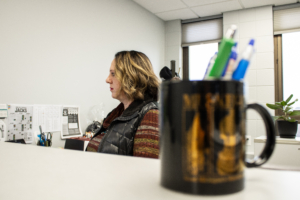 A woman with curly hair sits at a desk in an office, focused on her computer. A black mug with pens is in the foreground, with a plant nearby.