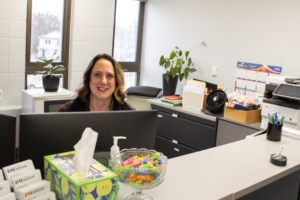A woman is smiling behind a reception desk with a computer. The desk has tissues, hand sanitizer, and a bowl of colorful candy. Plants and office supplies are in the background, creating a welcoming atmosphere.