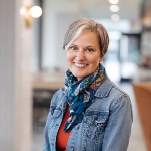 A woman with short gray hair smiles warmly, wearing a denim jacket, colorful scarf, and red shirt. The blurred background suggests a modern interior space.