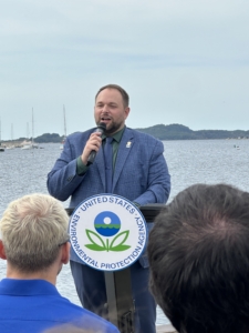 Mayor Ken Johnson speaks at a podium with the U.S. Environmental Protection Agency logo. He stands by the Muskegon Lake waterfront with boats and distant hills in the background.