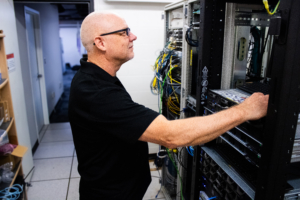 A man in a black shirt works attentively on a server rack, surrounded by colorful cables in a data center. The scene conveys focus and technical expertise.
