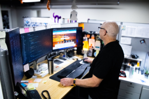 A man stands at a desk in an office, working at dual monitors displaying code and a website. The space is organized, with sticky notes and a lava lamp.