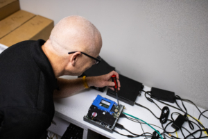A person with glasses and a bald head is fixing a circuit board on a white desk using a screwdriver. Wires and electronic devices are scattered around.