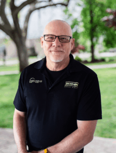 A bald man with glasses smiles warmly, standing outdoors. He wears a black polo shirt with logos. Green trees and a park setting are in the background.
