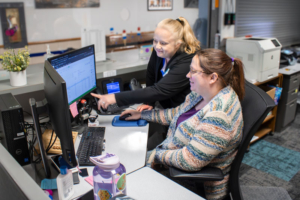 Two women at a desk, one seated and one standing, focused on a computer screen with spreadsheets. The atmosphere is collaborative and professional.