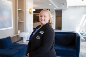A smiling person in a black jacket stands in a modern, bright lobby with plush blue sofas, abstract art, and soft lighting, conveying a welcoming tone.