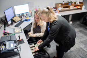 Two women are counting cash at an office desk. One is seated holding money, and the other is standing, placing bills in a drawer. Two monitors and office supplies are on the desk. The scene conveys a business or financial setting.