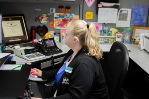 A woman with a ponytail sits at an office desk, focused on her computer. The desk is decorated with colorful artwork and personal items, creating a lively atmosphere.