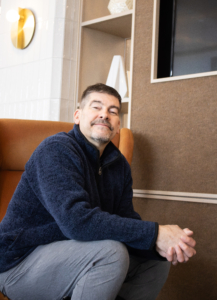 Man in a cozy setting sits on a brown chair, wearing a navy sweater. He appears relaxed and content, with a subtle smile. Neutral-toned decor.