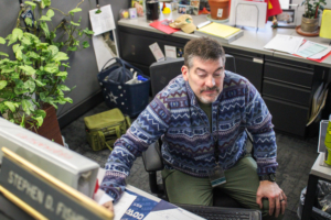 A man in a patterned sweater sits at a cluttered desk, reaching for a computer mouse. Office supplies and plants are visible. The atmosphere is busy yet focused.