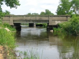 A stone bridge spans a calm, narrow river, surrounded by lush greenery. The scene is peaceful with a bright, clear sky above.