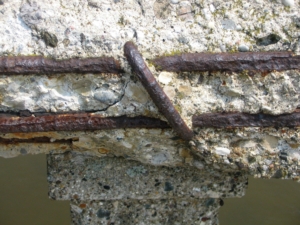 Close-up of a cracked concrete surface, revealing rusted steel reinforcement rods. The texture is rough, with small stones visible, conveying decay.
