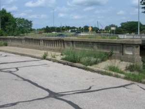 An old concrete bridge with overgrown grass and cracks on the pavement. Background shows a road with traffic signs and cars, under a cloudy sky.